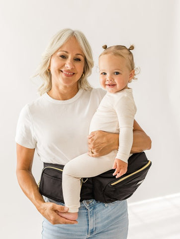 Caregiver carrying baby on their hip in a side-carrying position using a Tushbaby Hip Carrier, showing balanced support and comfort.